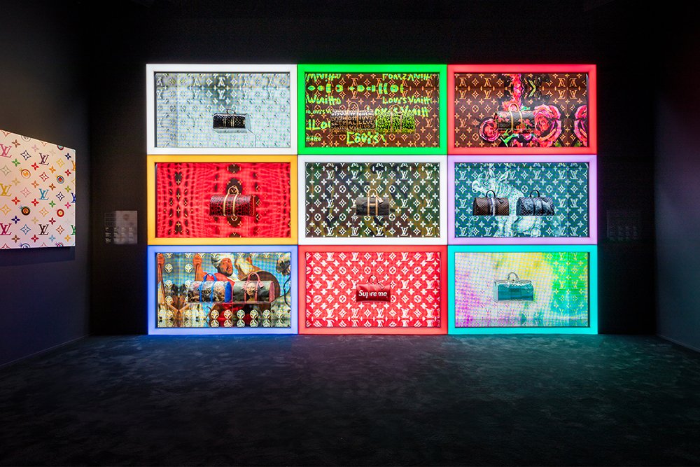 Dark gallery wall of nine neon-framed lightboxes displaying patterned bags and monogram artworks.