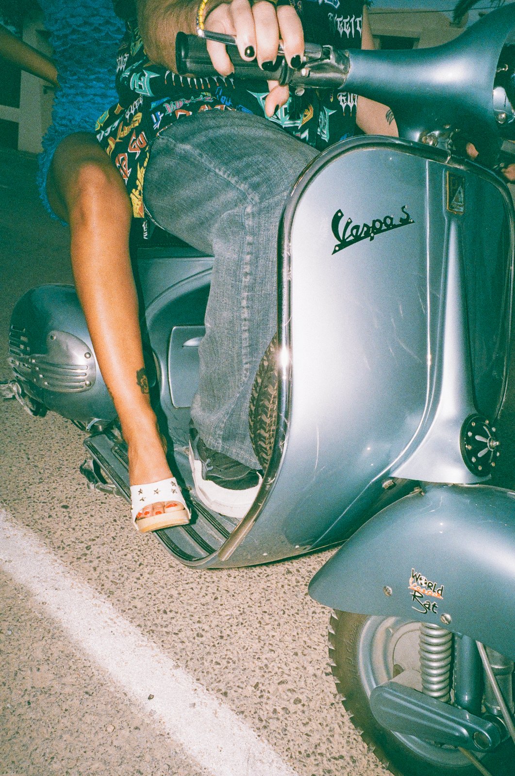 Playa Paradiso film still: a night-flash close-up of a woman's tanned leg in a starred white mule sandal resting on the running board of a pale-blue vintage Vespa, a man in a graffiti-print shirt gripping the handlebar.