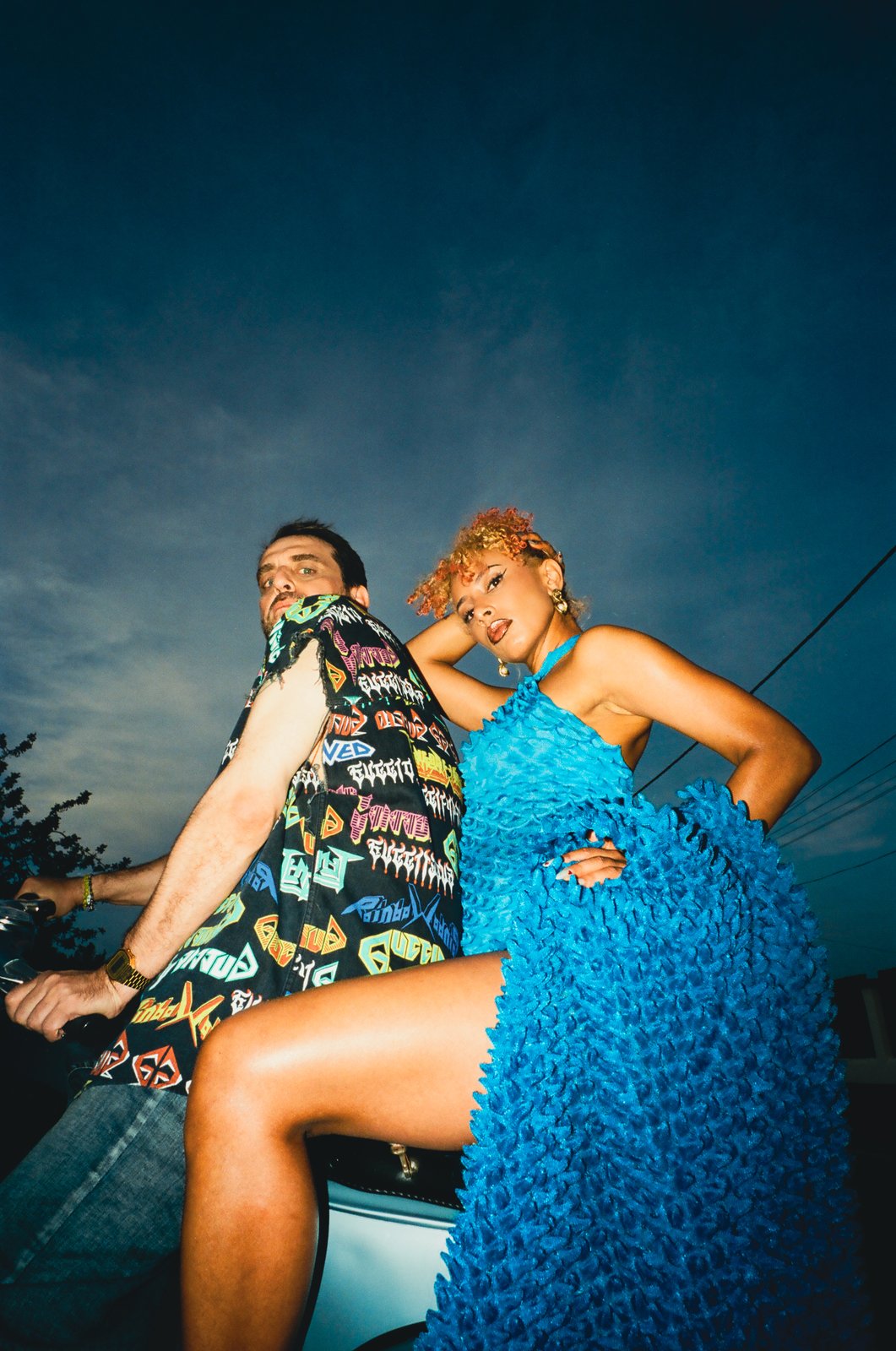 Playa Paradiso film still: a low-angle dusk portrait against a deep-blue sky of a man in a graffiti-print shirt and a woman with cropped orange curls in a bright-blue ruffled halter dress leaning back-to-back on the Vespa.
