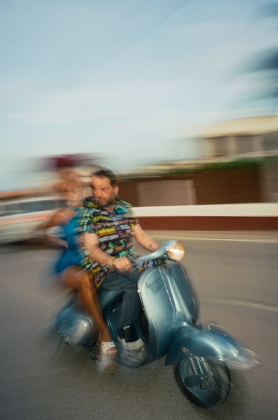 Playa Paradiso film still: a motion-blurred shot of the couple riding the pale-blue vintage Vespa along a seafront road at dusk, the man steering and the woman riding pillion in a blue dress.