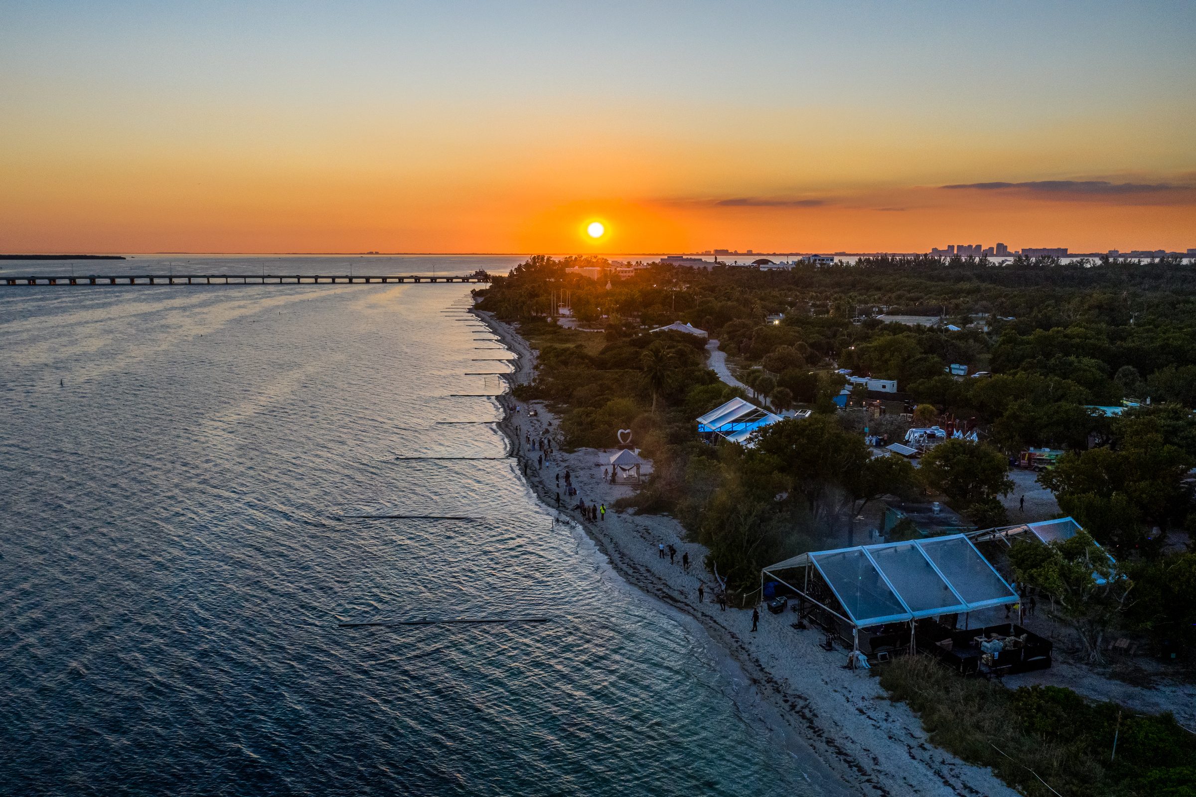 Aerial view at sunset of the mangrove coastline on Miami's Virginia Key, with festival marquees and a heart sculpture along the beach and the sun low over the bay.