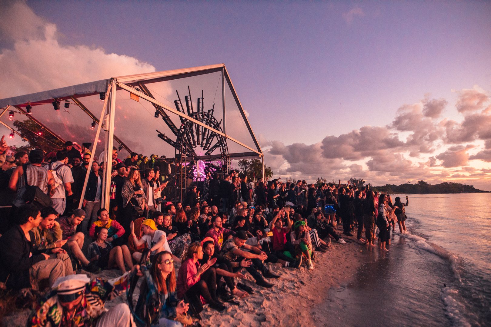 A dusk crowd gathered before the open-air festival stage and site installation, palms silhouetted against a fading sky.