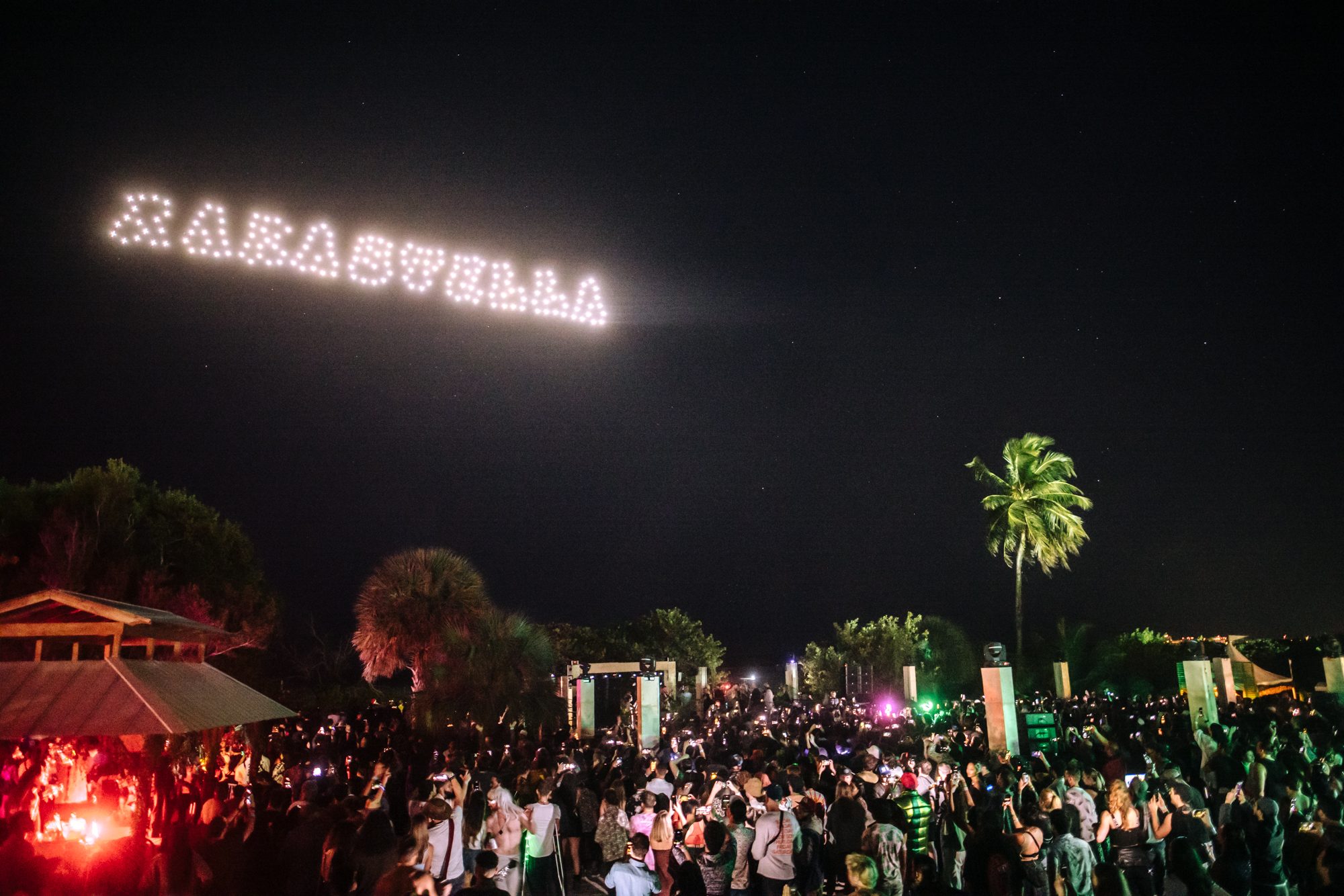 Hundreds of drones spelling “RAKASTELLA” in light across the night sky above a phone-lit crowd and palm trees.